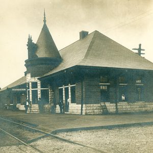 Single-story railroad depot building with conical tower and train tracks