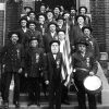 Group of older white men in matching uniforms with flag and drum on steps of brick building