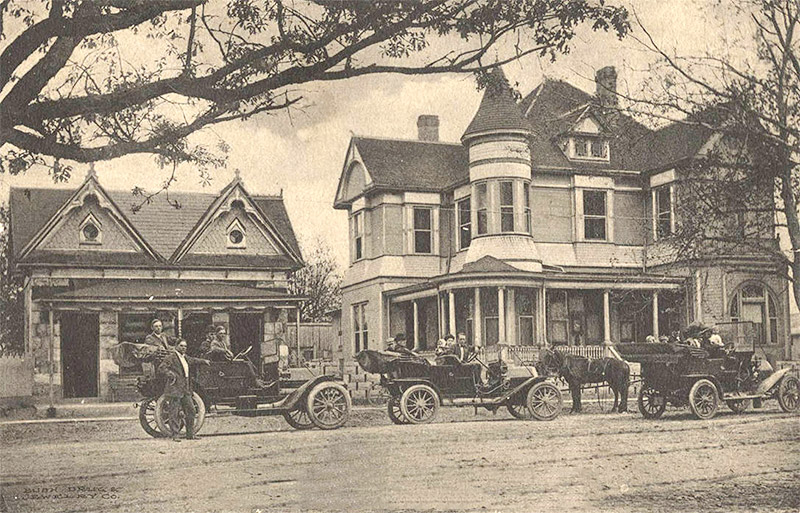 Men and women posing with three cars and horse on street outside single-story and multistory houses
