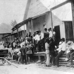 Crowd of people and line of buildings with a wagon on a dirt road