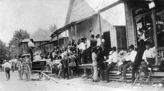Crowd of people and line of buildings with a wagon on a dirt road