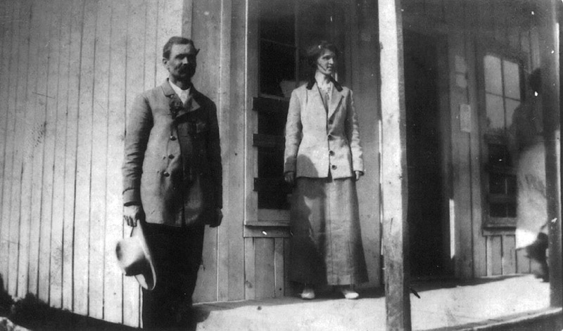 White man and white woman standing on before wooden building