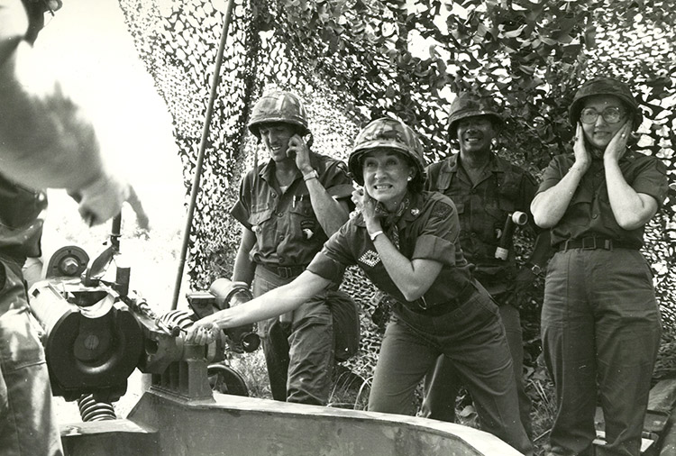 White woman in military uniform firing artillery from under canopy with other soldiers