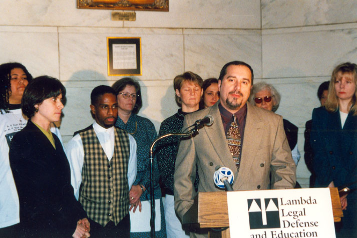 White man in suit speaking at a lectern with mixed group of men and women behind him