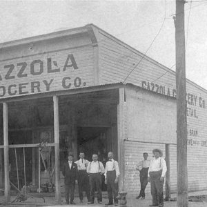 Group of white men standing outside "Gazzola Grocery Company" building on street corner