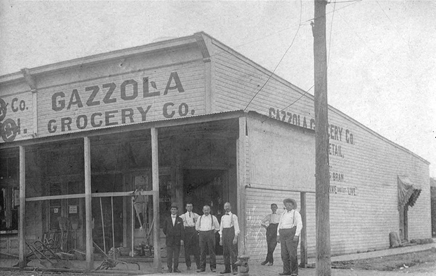 Group of white men standing outside "Gazzola Grocery Company" building on street corner