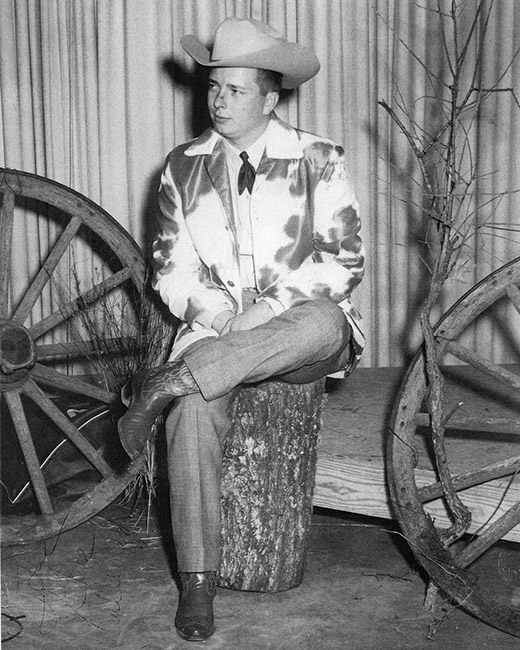 White man in cowboy hat and cowhide suit jacket sitting on prop stump between two wagon wheels and tree branches
