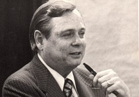 White man in suit and tie holding a pen