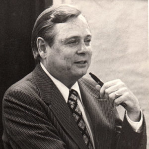 White man in suit and tie holding a pen