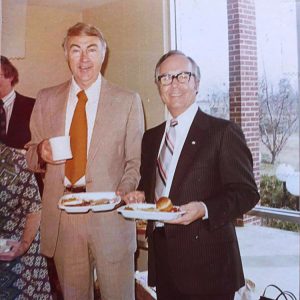 Older white man with coffee cup and tray standing next to white man in suit with glasses and tray