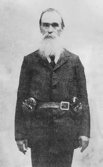 Older white man with long white beard standing in suit with hoster belt over his coat