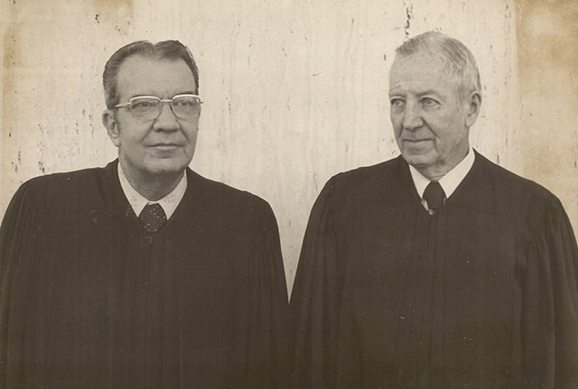 Older white man with glasses standing next to older white man with both wearing judge's robes