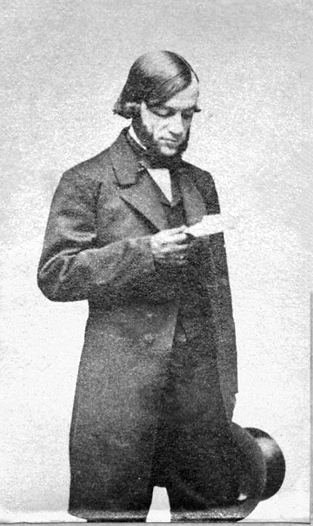 White man with sideburns in suit and bow tie reading a piece of paper