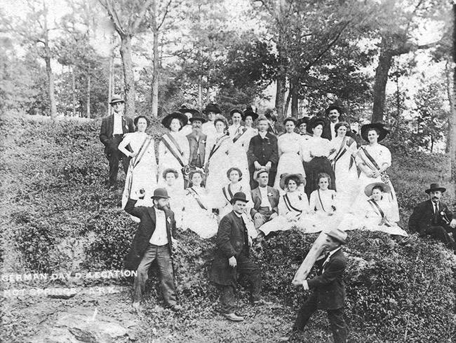 Group of white men in dark suits and women in white dresses with striped sashes on hill