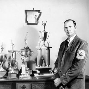 White man in suit with row of trophies and Arkansas plaque on the wall behind him