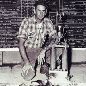 White man in flannel and hat kneeling with trophy and pile of fish with chalkboard behind him