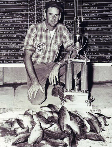 White man in flannel and hat kneeling with trophy and pile of fish with chalkboard behind him