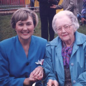 White woman smiling in blue seated next to older white woman with glasses smiling in blue coat