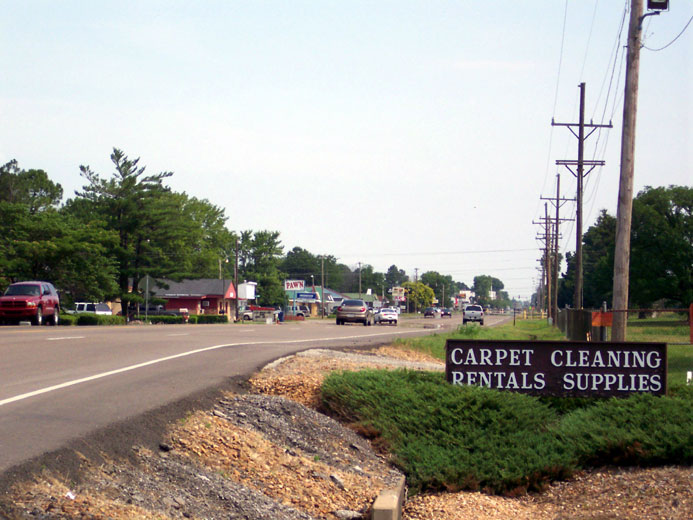 Street with cars and buildings on the left and "Carpet Cleaning Rental Supplies" sign next to a line of telephone polls on the right