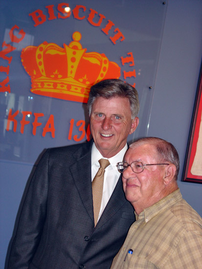 Older white man in suit and tie standing next to older white man in plaid shirt with crown "King Biscuit Time" logo on the wall behind them