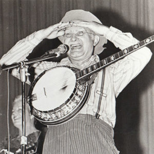 White man at microphone with banjo, shading eyes