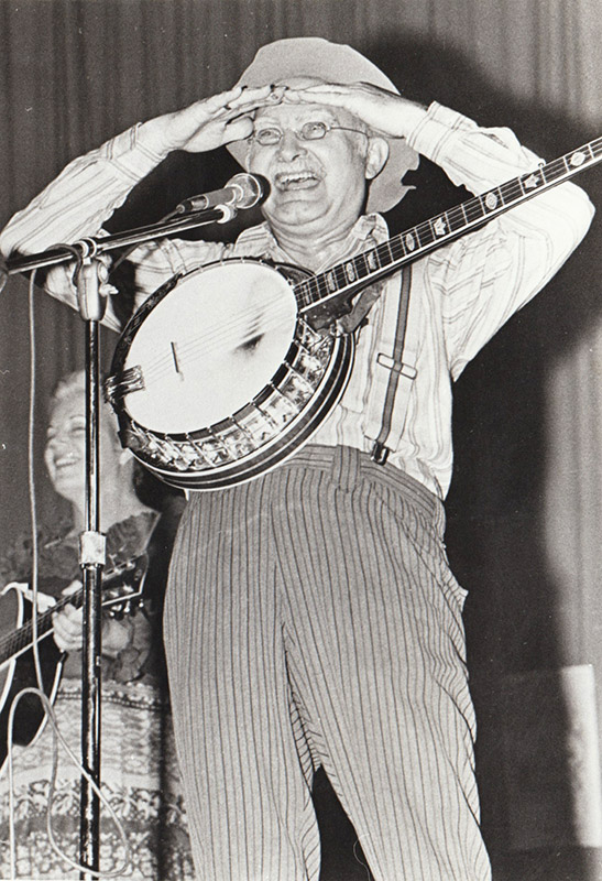 White man at microphone with banjo, shading eyes