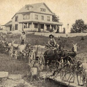 White men and children with horse and mule drawn wagons and multistory house in the background