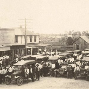 Crowd of white men and cars on town street with storefronts