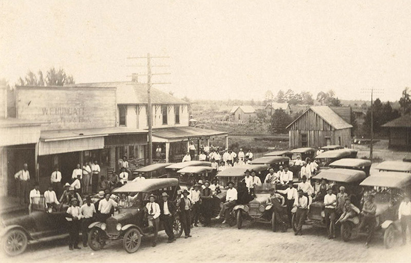 Crowd of white men and cars on town street with storefronts