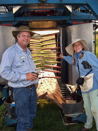 White man in hat and denim uniform and woman in hat and blue shirt standing inside grape harvesting machine