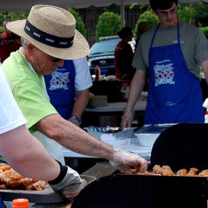 Older white man in hat and white men in blue aprons grilling  outdoors