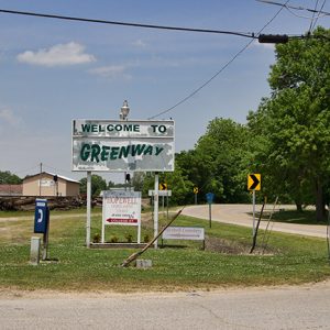 "Welcome to Greenway" sign and road signs on rural road with single-story buildings in the background