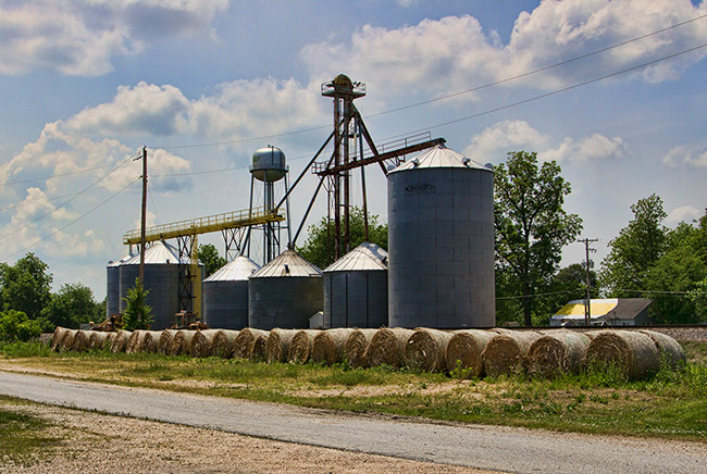 Row of hay bales in front of grain elevator and silos with water tower in the background