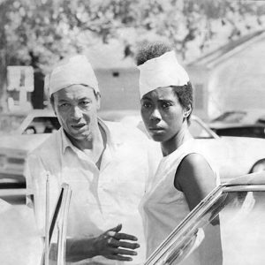 African American man and woman standing with cars and buildings behind them