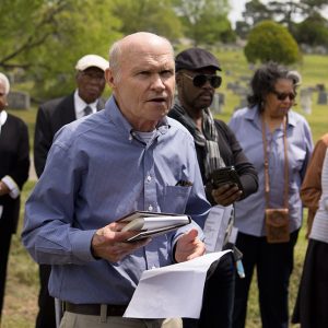 Old white man holding book and notes speaking with African-American men and women behind him