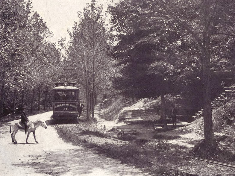Man on horseback crossing in front of trolley car on tree-lined street with man standing under trees on the other side