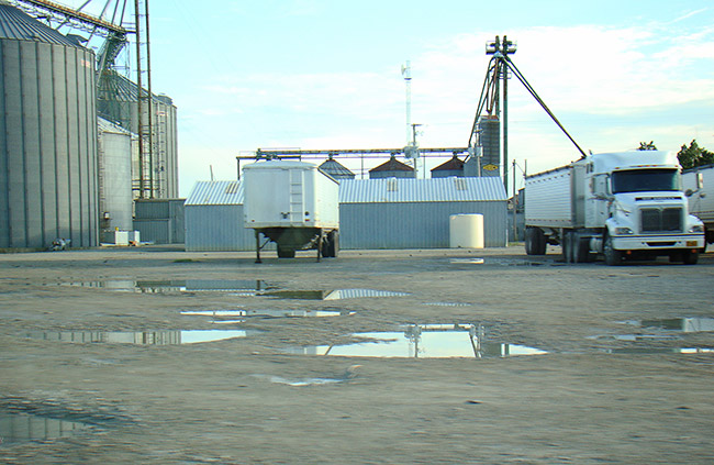 Semi-trucks and trailer parked outside large silos on parking lot with puddles