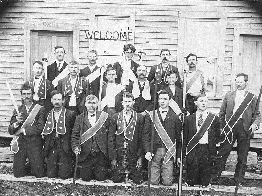 Group of white men in suits with different types of ribbons and sashes poses outside building