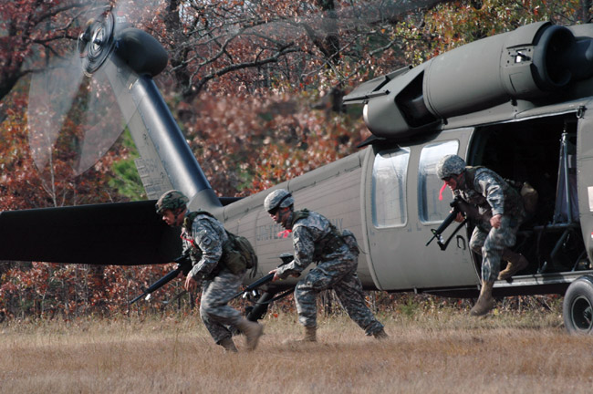 Three armed male soldiers hurriedly exiting powered helicopter in field near tree line
