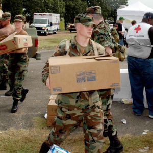 White men and women soldiers unload boxes from truck near American Red Cross volunteers and crowd