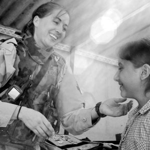 White woman soldier physician inspects young girl in blouse ponytail both smiling indoors