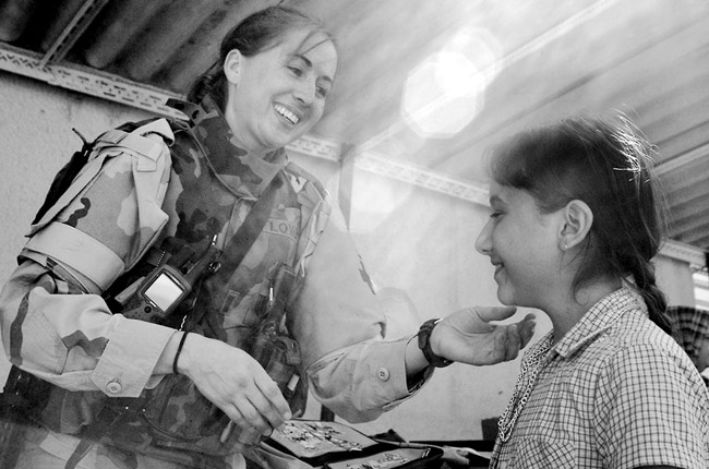 White woman soldier physician inspects young girl in blouse ponytail both smiling indoors