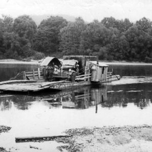 man and truck on ferry boat crossing a river with trees in background