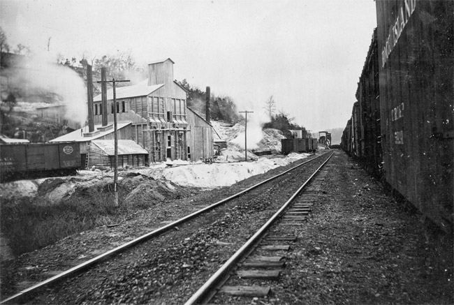 Railroad track running next to a two-story mining building with two smoke stacks and power lines