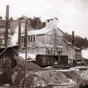 Multistory industrial complex featuring multiple smokestacks perched on a hill with railcar in front
