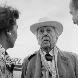 White man and woman greet older white man in hat and suit with airplane in the background