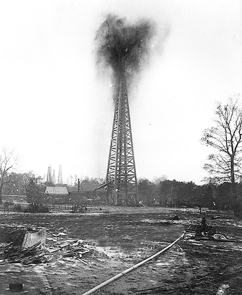 Oil gushing from a steel derrick with single-story building nearby