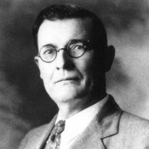 Portrait of white man with short combed hair with suit jacket tie round glasses and serious expression