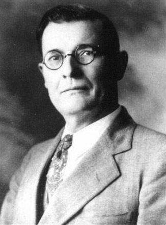 Portrait of white man with short combed hair with suit jacket tie round glasses and serious expression