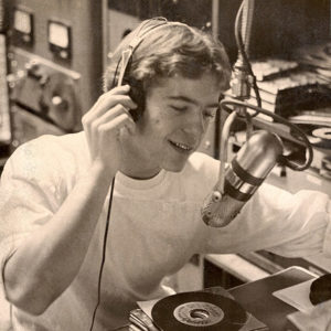 Young white man with headphones speaking into a microphone in radio booth
